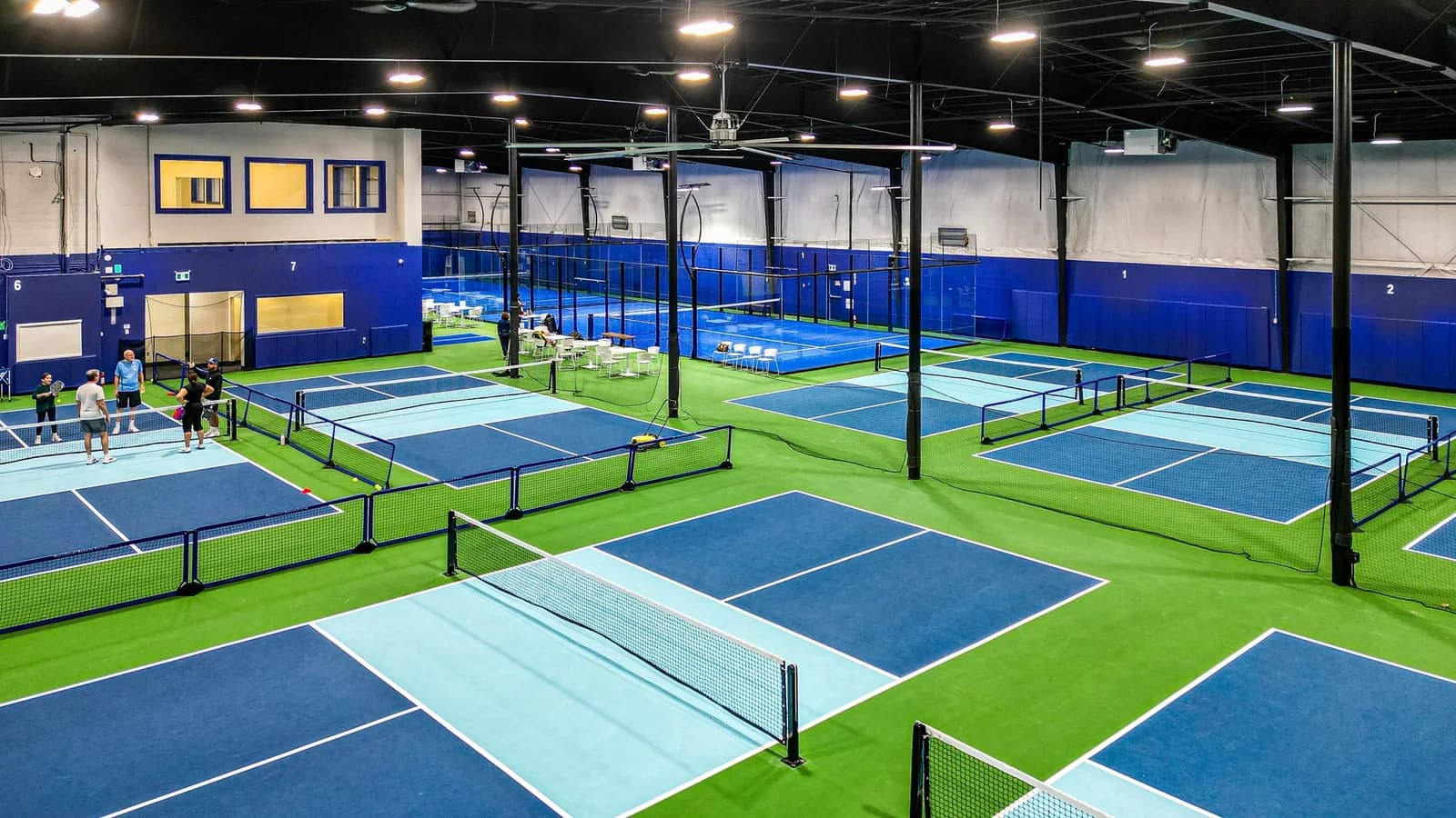 Elevated view of an indoor pickleball facility with rows of blue and green courts under high industrial ceilings