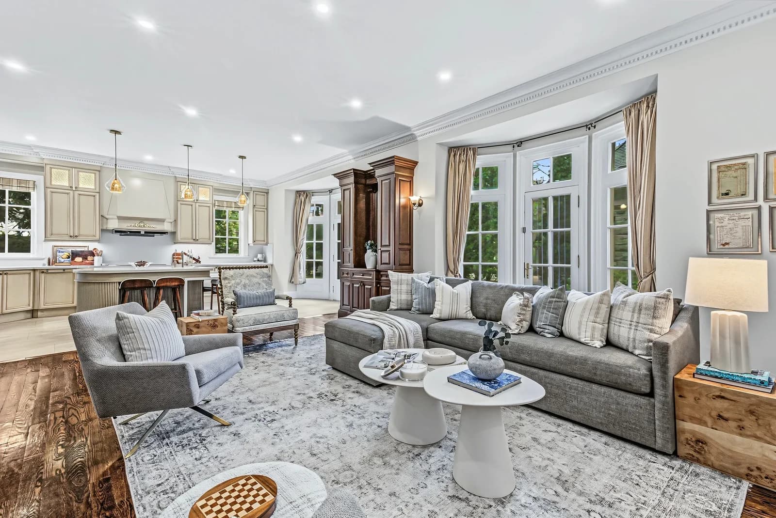 Bright open-concept living room with a grey sectional, round white coffee tables and bay windows, opening to a cream kitchen with pendant lights