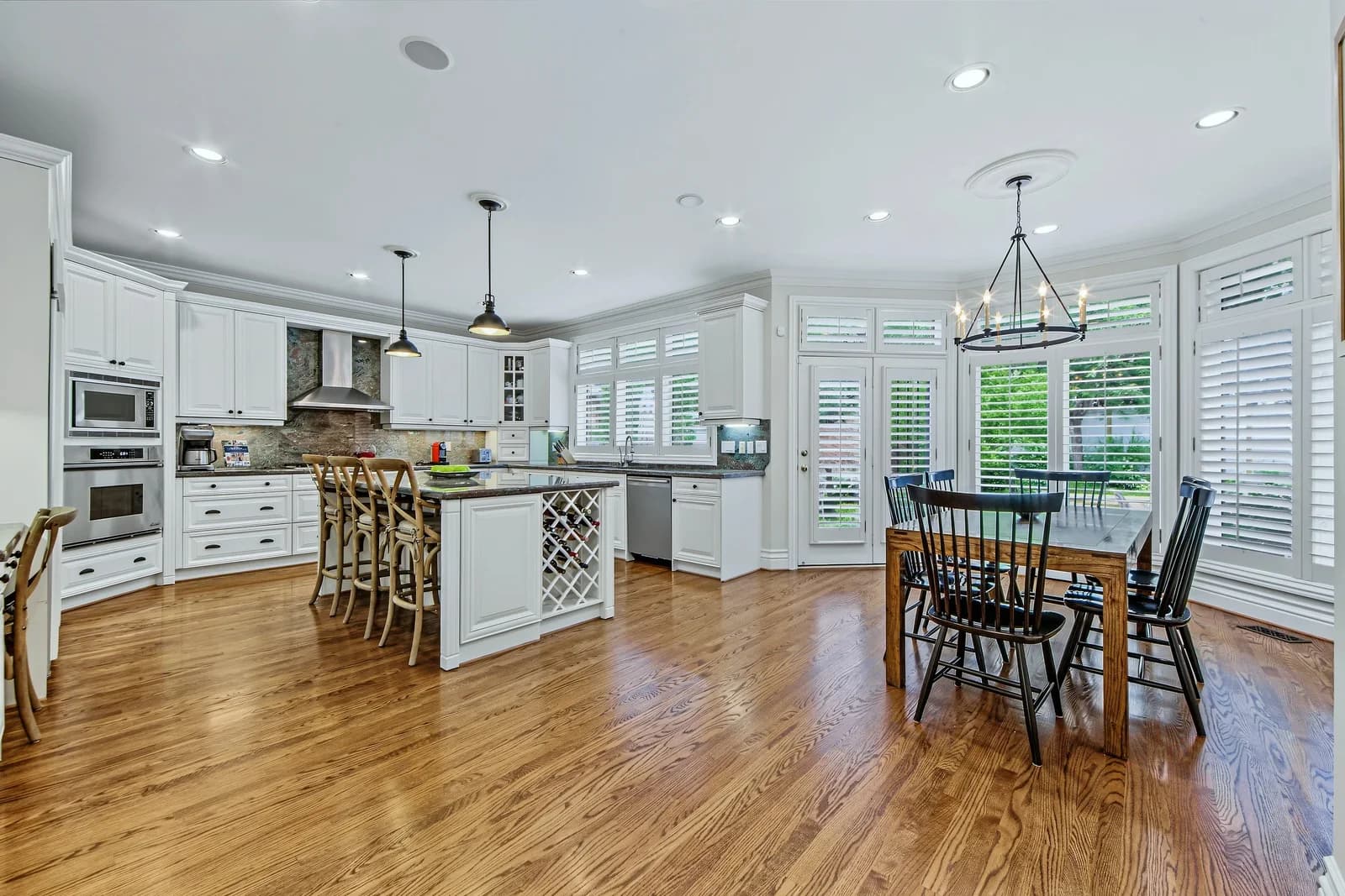 White kitchen with a large island, stainless appliances and hardwood floors, next to a breakfast area with a wooden table under a black chandelier